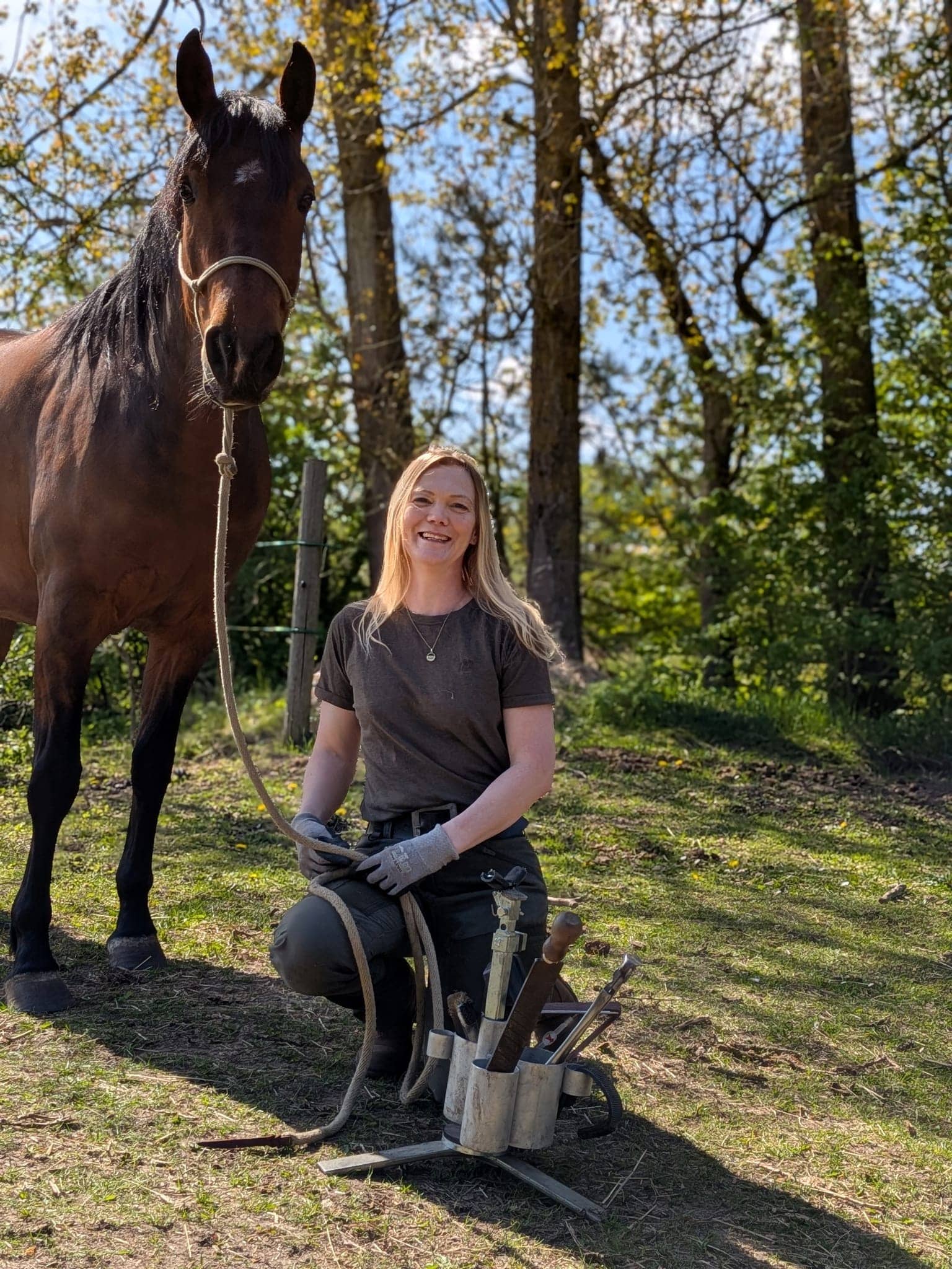 Marlene knæler smilende ved siden af en brun hest med sit værktøj